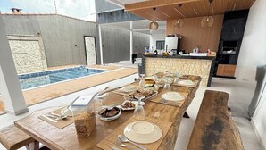 A long wooden dining table set with plates, cutlery, and glassware, with a view of a swimming pool and a modern kitchen in the background. - MC Style house in Salinas, PA, 8 beds and 4 suites (Salinópolis)