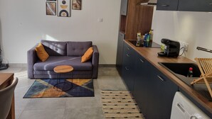 A modern kitchen with dark cabinets, a built-in oven, and a coffee machine. There is a gray sofa with orange cushions and a small wooden table. - Quiet apartment in a green setting (Nîmes)