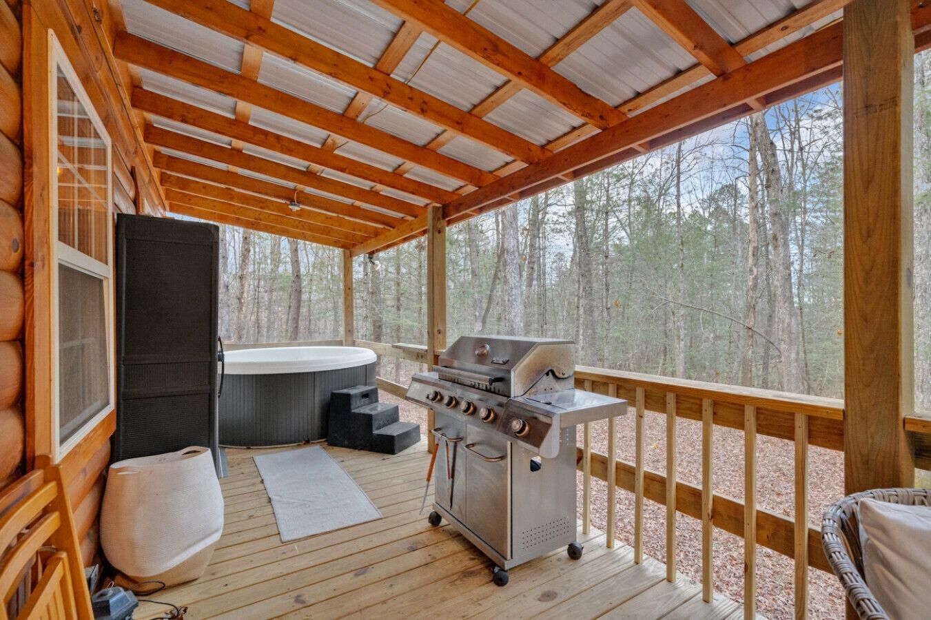 A covered outdoor deck with a hot tub, grill, and seating area, surrounded by a forest.