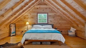 Un dormitorio acogedor en el ático con techo inclinado de madera, una cama con colcha azul y blanca, una mesita de noche con lámpara y una ventana con vistas al verdor.