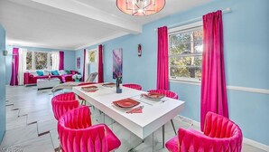 Un coin repas avec une table blanche dressée pour quatre personnes, des chaises roses et un lustre.