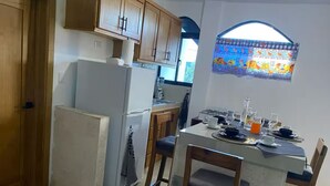 Une cuisine avec des meubles de rangement en bois, un réfrigérateur, une table à manger pour quatre personnes et une fenêtre ornée d’un motif coloré.
