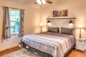 Un dormitorio con una cama, dos lámparas de noche, una mesita de noche, una ventana con cortinas florales, un ventilador de techo y un cuadro en la pared.
