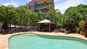 Un área de piscina con un pabellón, sillones de descanso y un edificio de varios pisos al fondo.