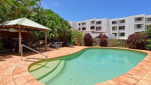 Una piscina con plataforma de ladrillo, un área de estar techada con sillas, y un edificio al fondo.