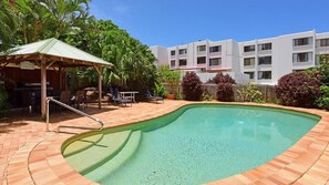 Una piscina con plataforma de ladrillo, un área de estar techada con sillas, y un edificio al fondo.