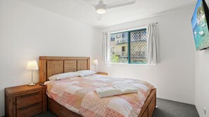 A bedroom with a wooden bed, two bedside lamps, a window with a view of buildings, and a ceiling fan. - Lindomare Unit 10, 11 Orvieto Tce, Kings Beach (Kings Beach)