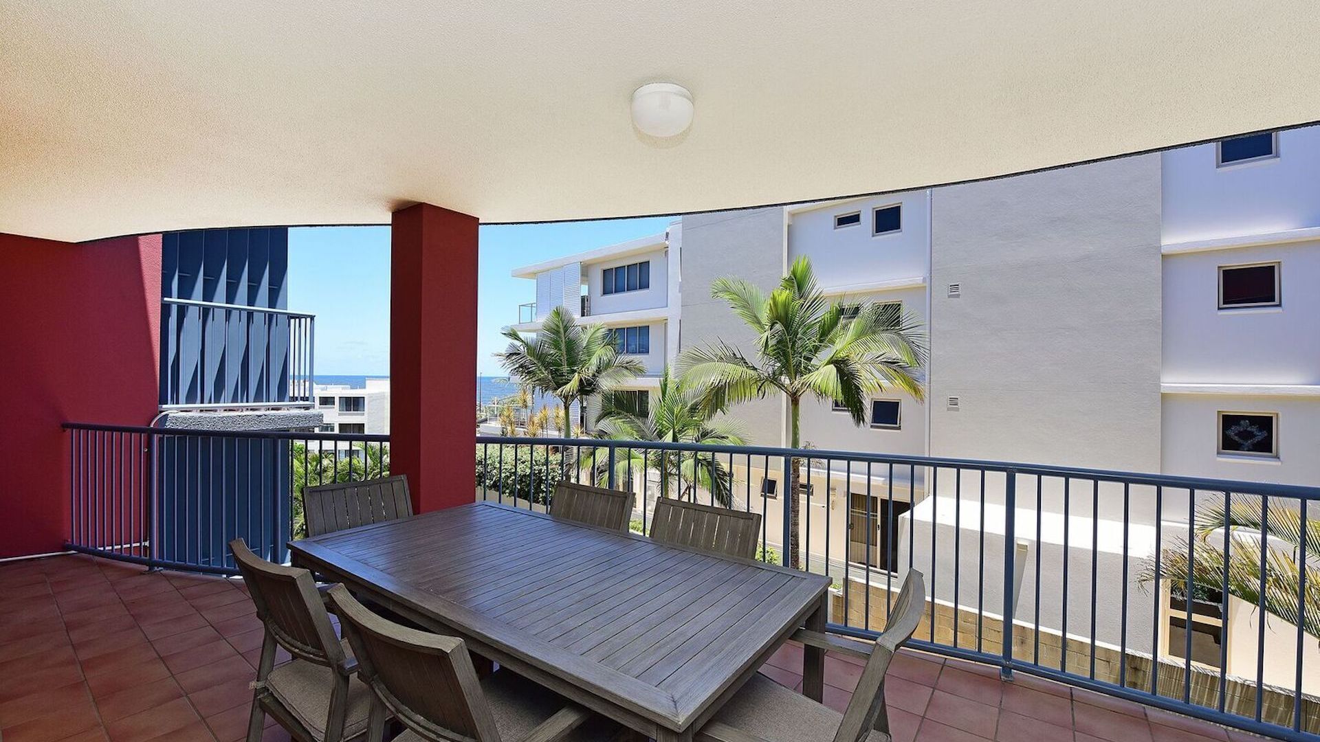 A balcony with a wooden table and chairs, a grill, and a view of palm trees and buildings.