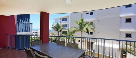 A balcony with a wooden table and chairs, a grill, and a view of palm trees and buildings.