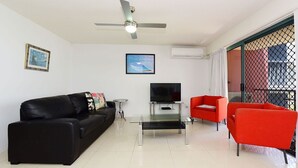 A living room with a black sofa, a red armchair, a glass coffee table, a flat-screen TV, and a ceiling fan. - Lindomare Unit 12, 11 Orvieto Tce, Kings Beach (Kings Beach)
