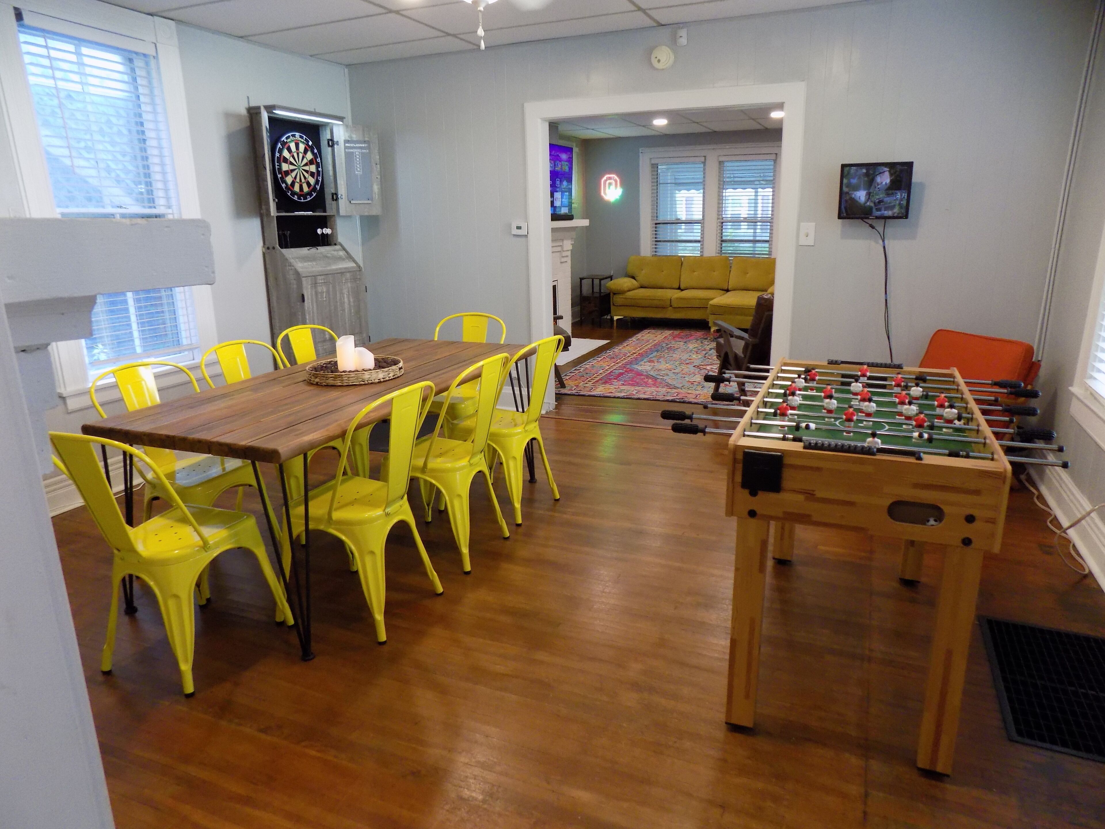 A room with a wooden table and yellow chairs, a dartboard, a foosball table, and a television on the wall.