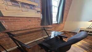 A room with a glass-top desk, a black office chair, a brick wall, and a window with curtains.