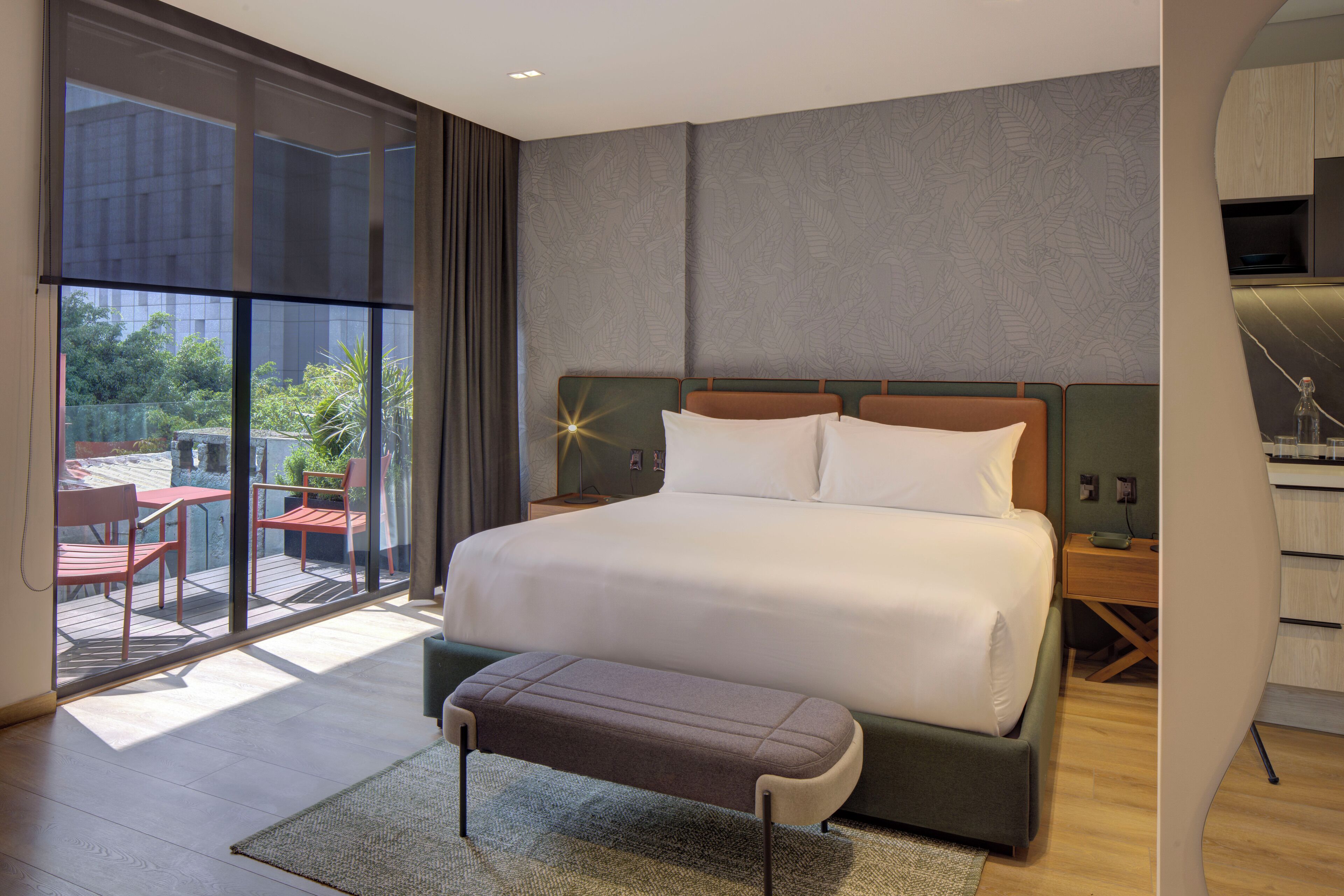 A modern hotel room with a large bed, a bench, a desk, and a view of the outdoors.