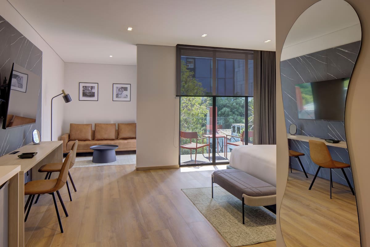 Una habitación de hotel moderna con sofá, una mesa de comedor, sillas, televisión y un balcón con mobiliario exterior.