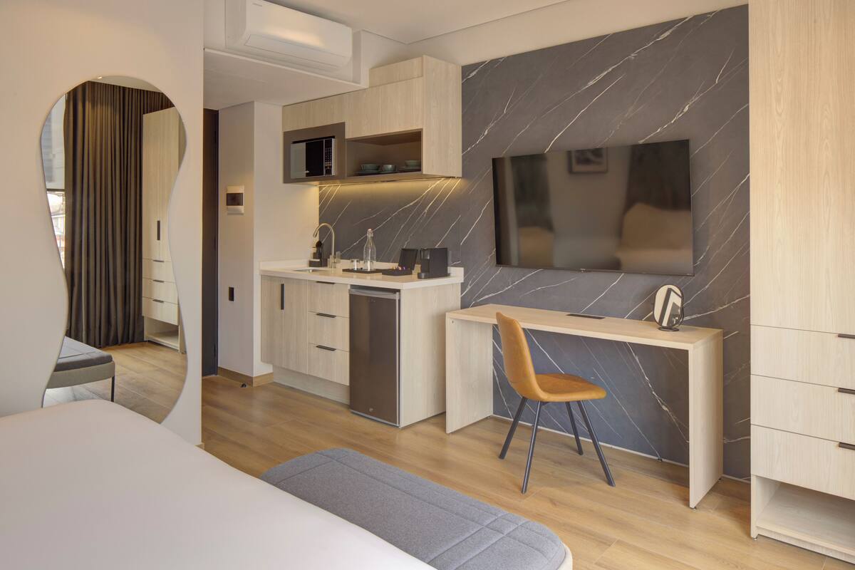 Una habitación de hotel moderna con una cama, un escritorio con una silla, una kitchenette pequeña y un espejo grande en la pared.