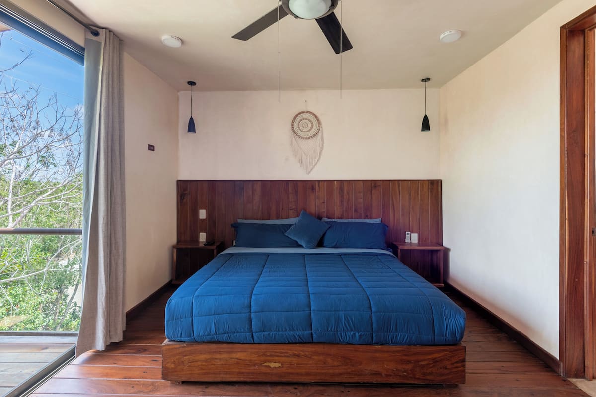 Un dormitorio con una cama grande, cabecera de madera, dos mesitas de noche, un ventilador de techo y vistas a través de un ventanal amplio.
