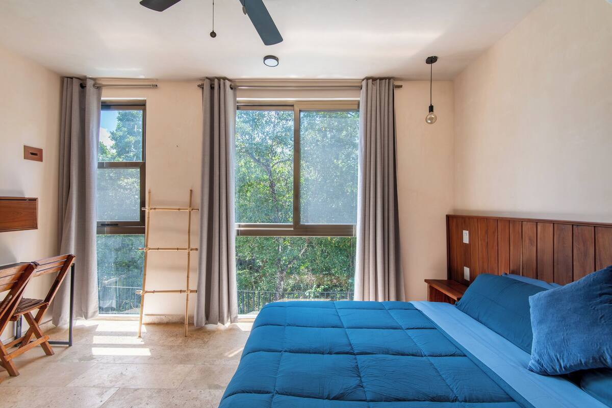 Un dormitorio con una cama grande, cabecera de madera, ventana con cortinas, ventilador de techo y un banco de madera.