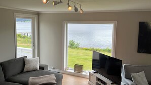 A living room with a large window overlooking a body of water, a gray sofa, a television on a stand, and ceiling lights. - Fabulous 4-bedroom villa with AC in Møre og Romsdal, boat available (Møre og Romsdal)