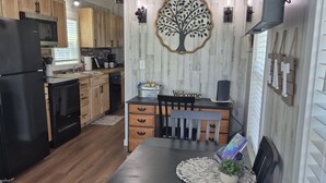 A modern kitchen with wooden cabinets, a black refrigerator, a dining table with chairs, and a wall-mounted light fixture. - Sunrise Haven Tiny Home (Big Cabin)