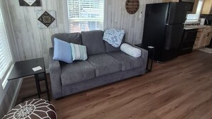 A modern living room with a grey sofa, a wooden floor, a ceiling fan, and a kitchen area in the background. - Sunrise Haven Tiny Home (Big Cabin)