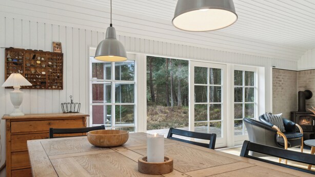 A wooden dining table with a candle, a bowl, and a lamp. There are chairs around the table, a fireplace, and a large window with a view of trees.