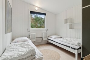 Une petite pièce aux murs blancs, avec deux lits, une fenêtre et une étagère fixée au mur.