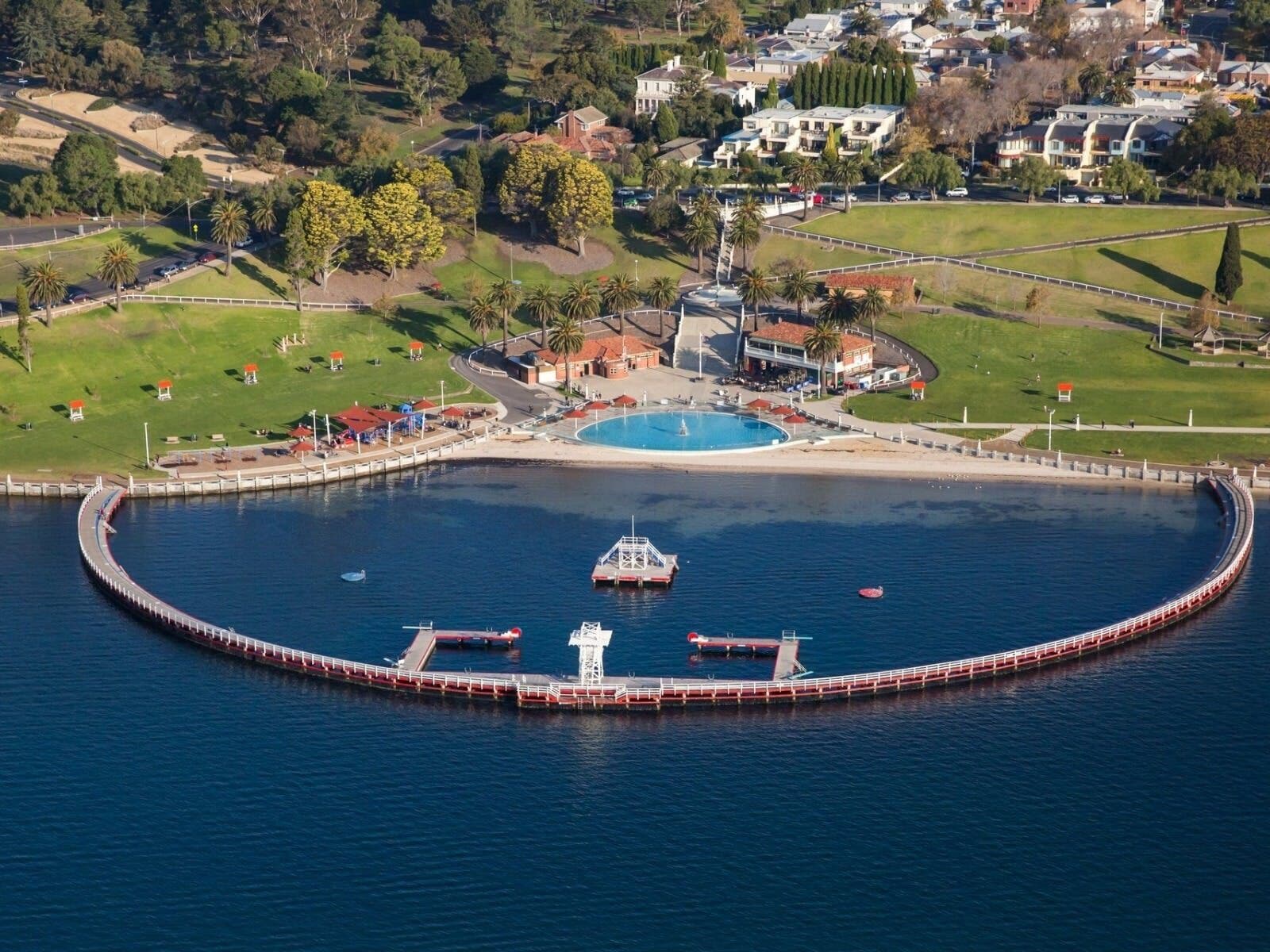 Vista aérea de uma área costeira com um porto circular, uma piscina e edifícios residenciais ao fundo.