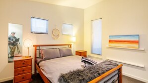 A bedroom with a bed, a wooden dresser, a desk with a lamp, a camera on a tripod, and a painting on the wall.