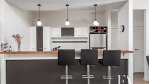 A modern kitchen with a central island, three bar stools, pendant lighting, and a clock on the wall.