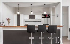 A modern kitchen with a central island, three bar stools, pendant lighting, and a clock on the wall.