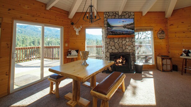 Ein Holzhütten-Interieur mit Kamin aus Stein, einem Holzesstisch mit Bänken, einem großen Fenster mit Blick auf einen Wald und einer Terrassenfläche.