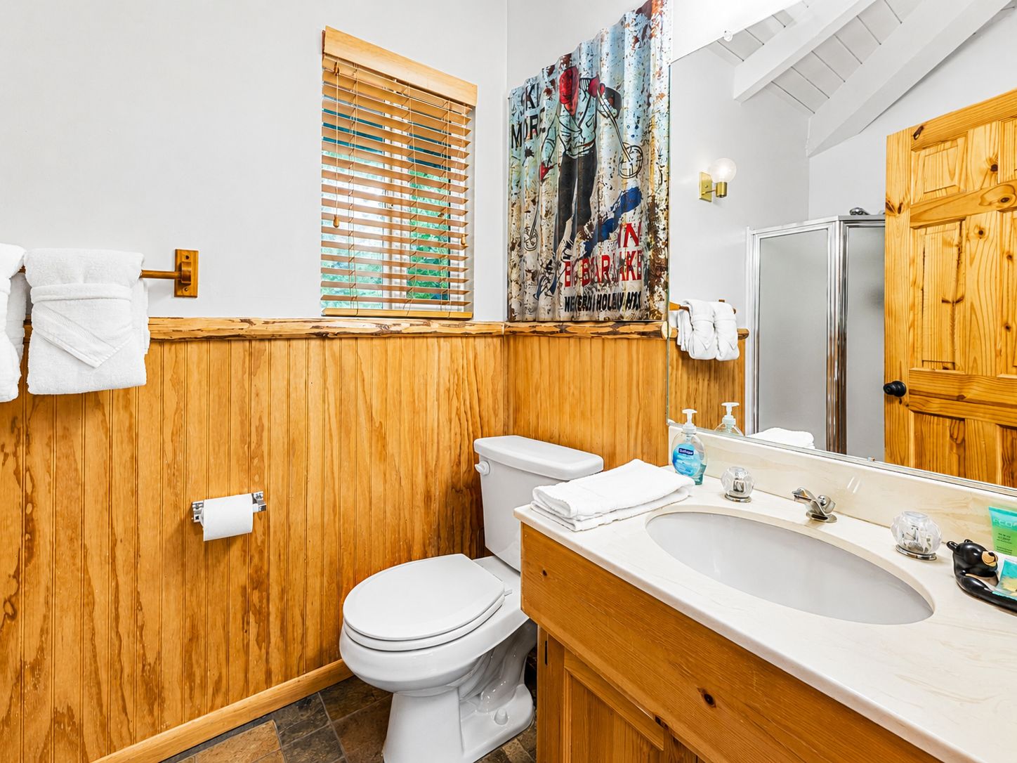 A bathroom with a wooden vanity, a toilet, a sink, and a window with blinds.