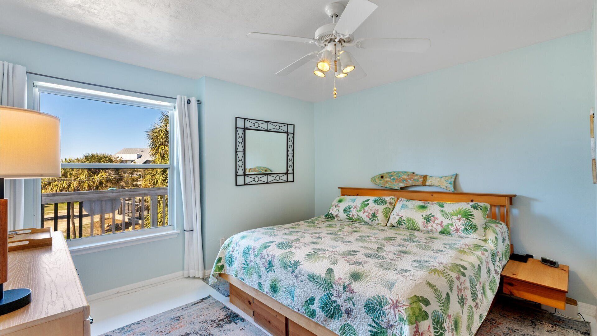 A bedroom with a bed, a ceiling fan, a window with a view, and a small desk with a lamp.
