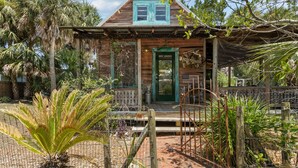 A rustic wooden house with a green door and windows, surrounded by palm trees and a fenced garden.