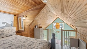 A bedroom with a wooden ceiling, a bed with a patterned duvet, a dresser, a mirror, and a view of the outside.