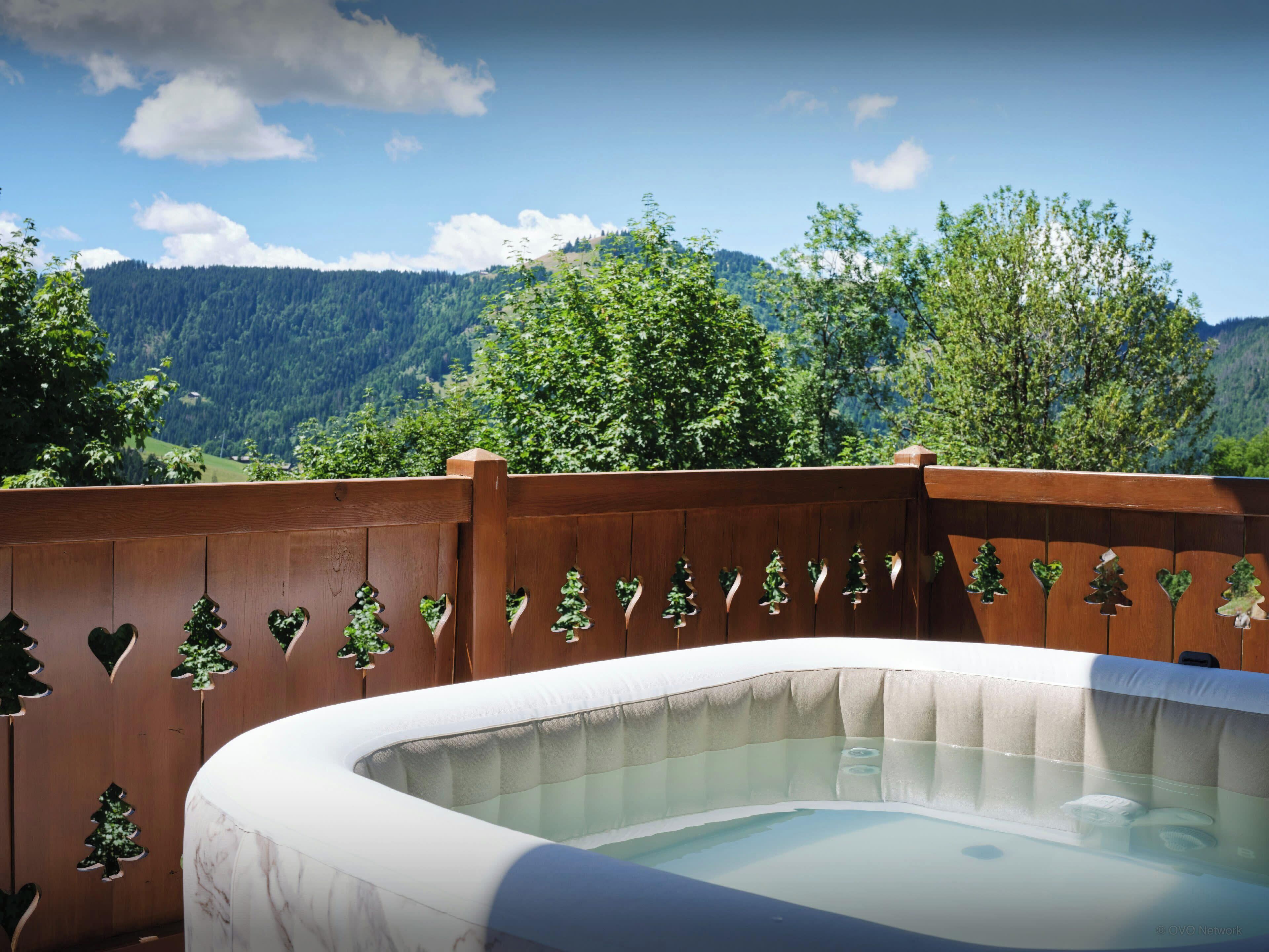 Un bain à remous entouré d’une clôture en bois ornée de motifs en forme de cœur, le tout se fondant dans un paysage montagneux.