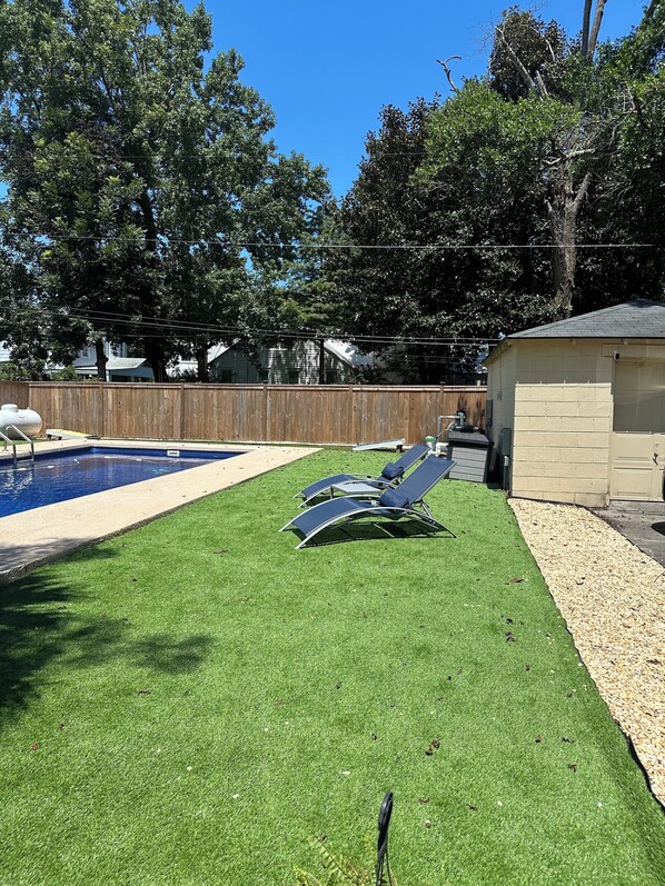 A backyard with a swimming pool, artificial grass, and lounge chairs.
