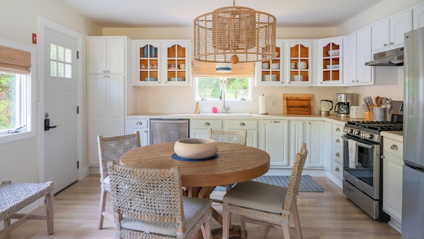 A modern kitchen with white cabinets, a round wooden table, wicker chairs, and a hanging light fixture.