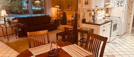 A dining area with a table set for four, a kitchen with white cabinets, and a view of the outdoors through a large window.