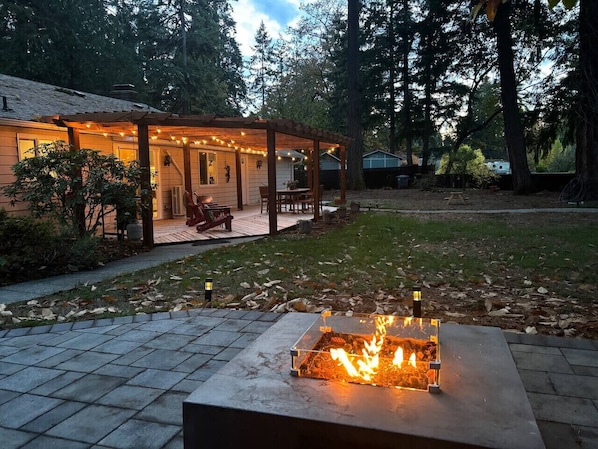 Una terraza de madera con un pozo de fuego y luces colgantes, rodeada de zona boscosa.