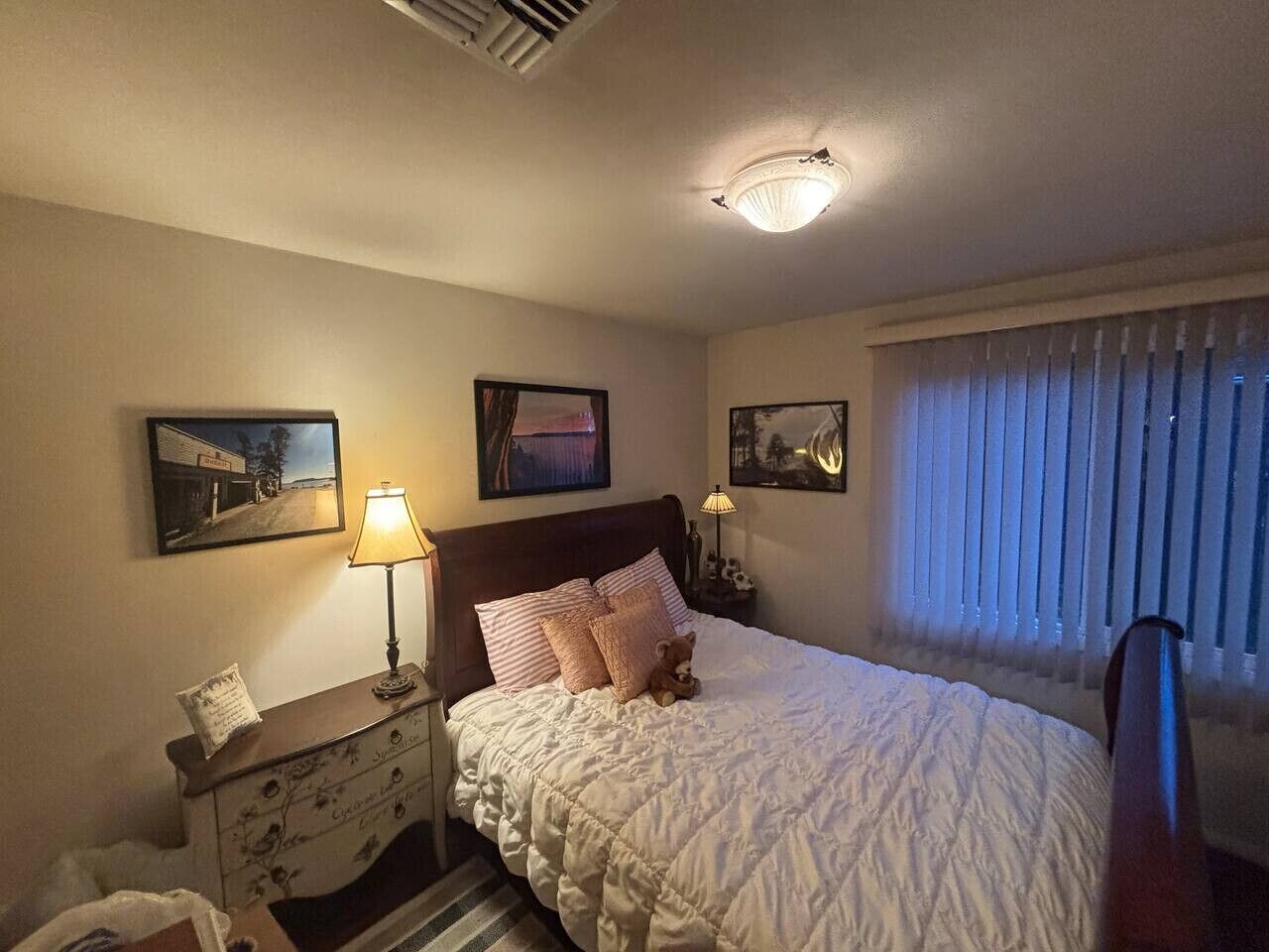 Un dormitorio con una cama, una mesita de noche, una lámpara, un aparador y dos cuadros en la pared.