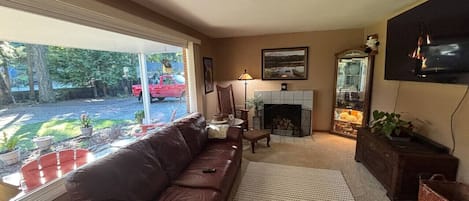 Una sala de estar con un sofá de cuero marrón, chimenea, televisor montado en la pared y vistas al exterior.