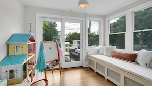 A bright room with a large window, a wooden floor, a white bench with cushions, and a dollhouse.