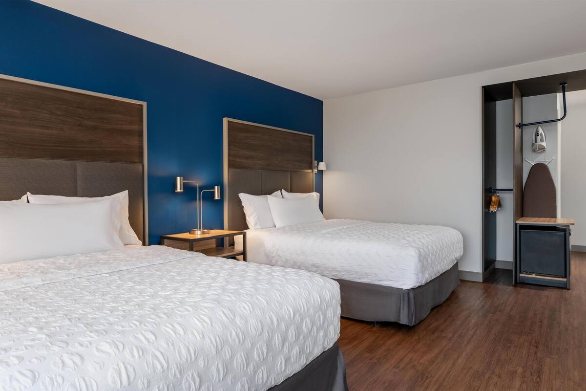 A hotel room with two beds, a blue accent wall, a wooden headboard, a nightstand, and a small table with an ironing board.
