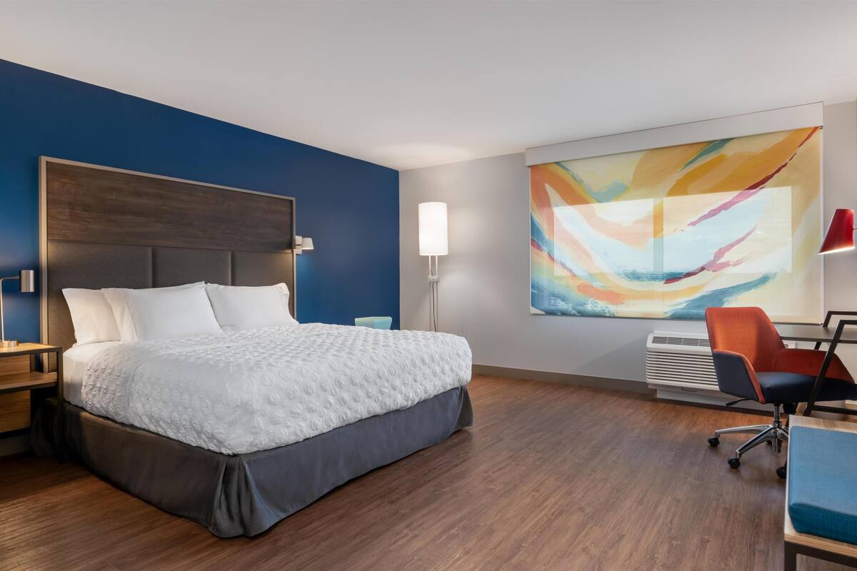 A modern hotel room with a large bed, a desk with a chair, a standing lamp, and a colorful abstract painting on the wall.