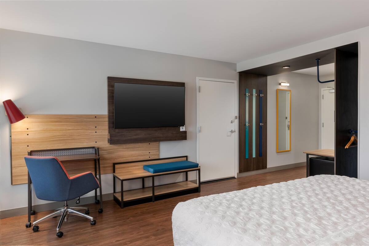 A modern hotel room with a bed, a desk with a chair, a television, and a wardrobe with a mirror.