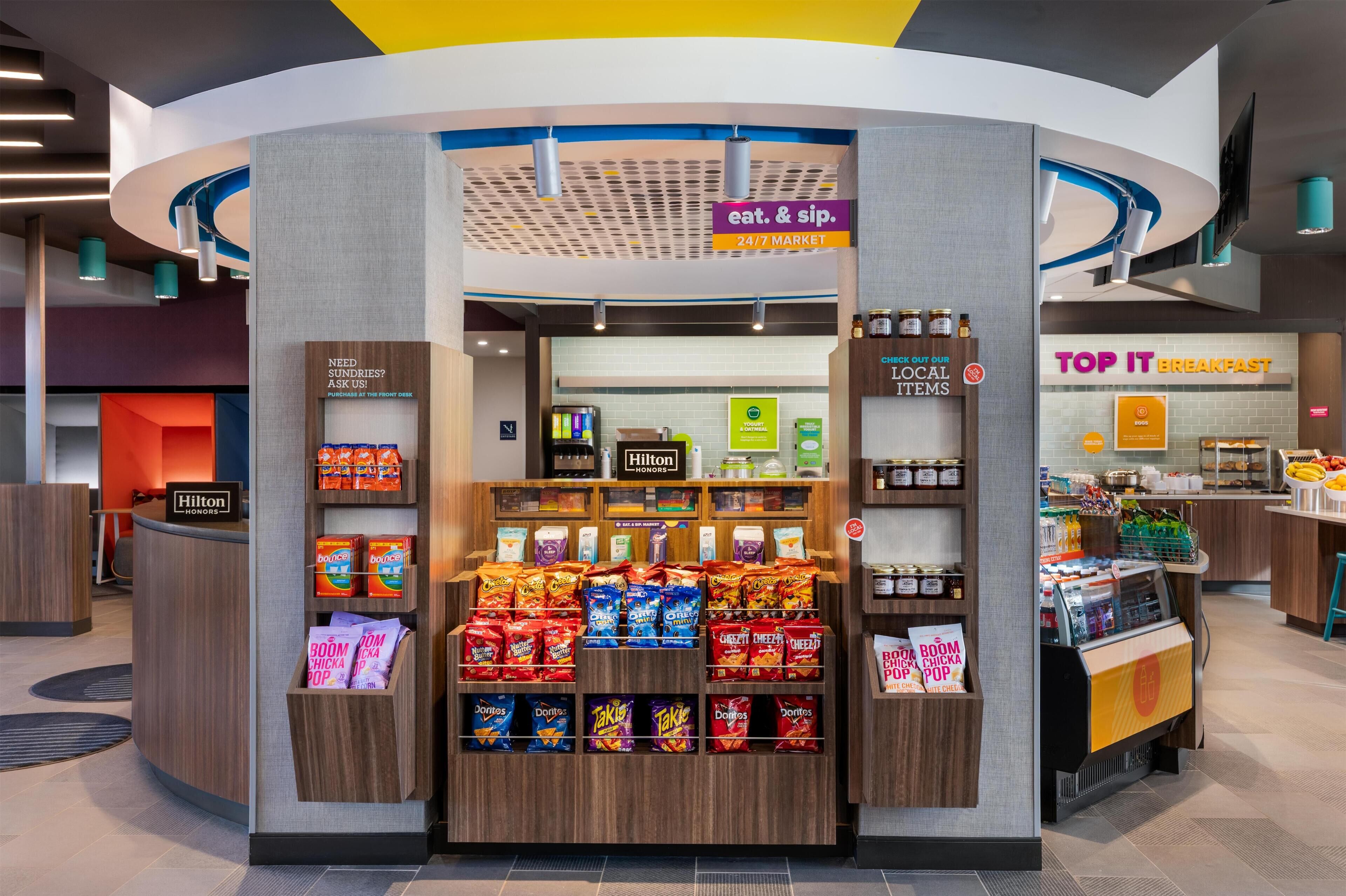 A snack kiosk with a variety of chips and snacks, a 24/7 market sign, and a breakfast counter with fresh produce.