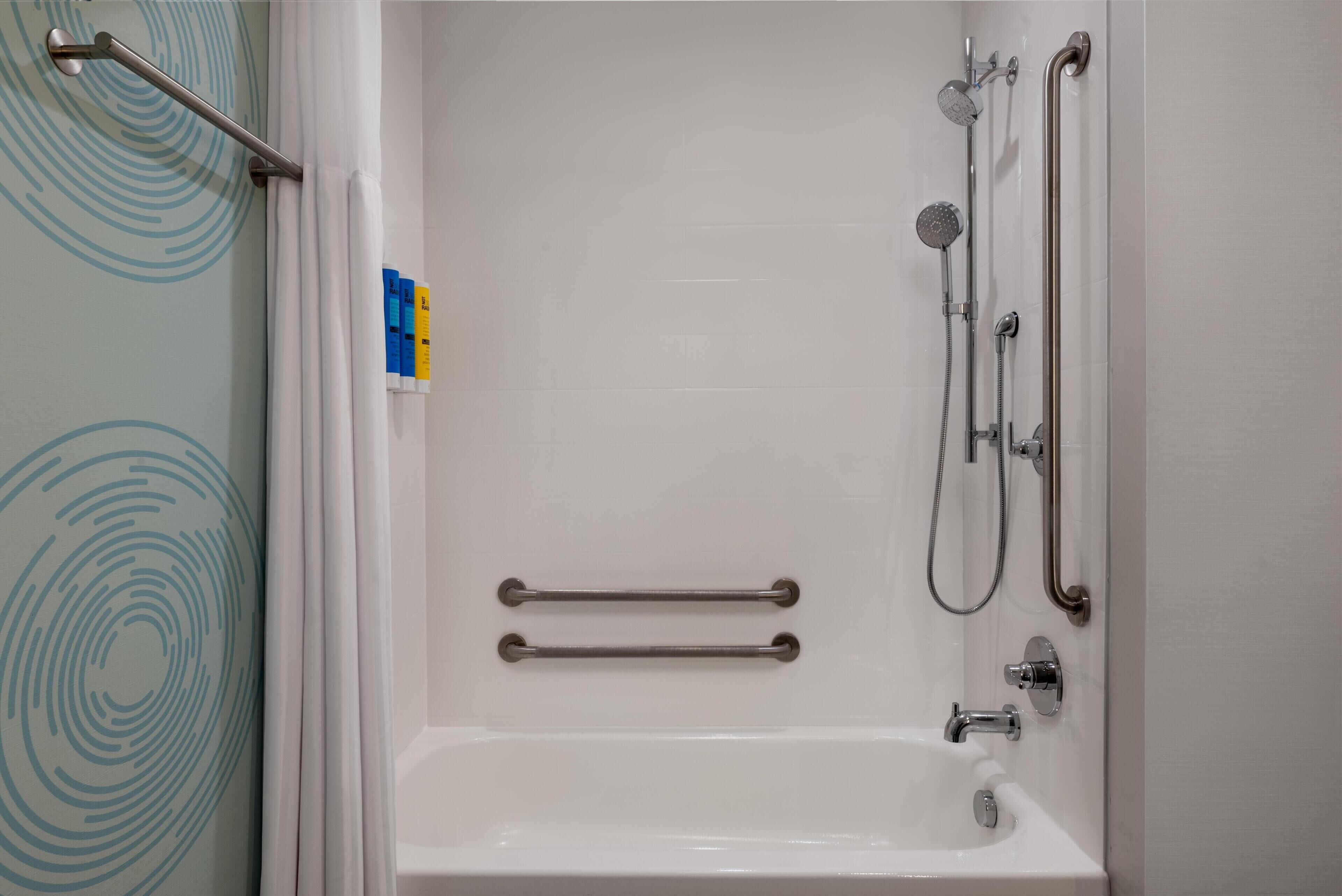 Un baño con bañera blanca, barras de apoyo, cabezal de ducha y ducha de mano.