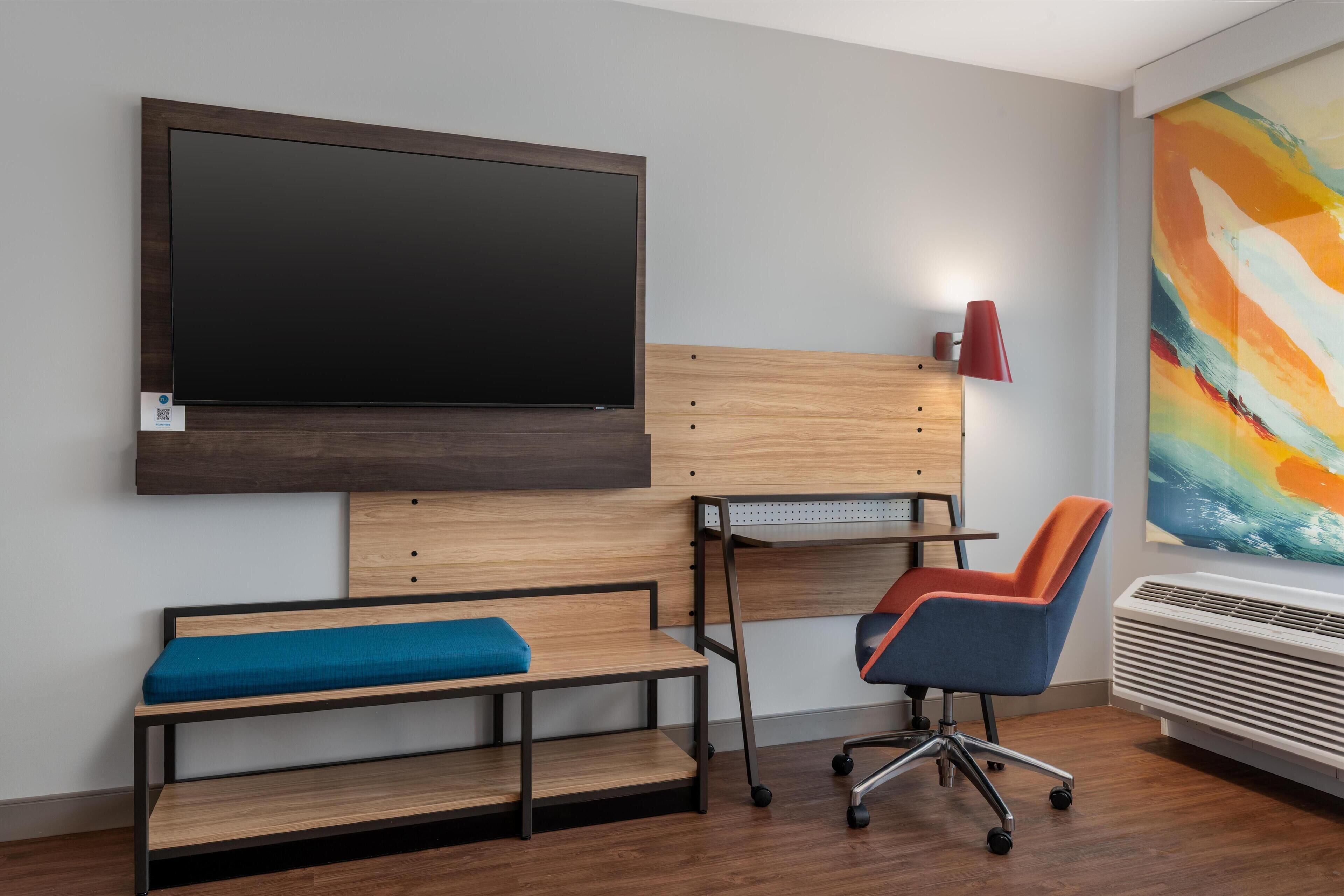 A modern room with a large flat-screen TV mounted on the wall, a wooden desk with a chair, a bench with a cushion, and a colorful abstract painting on the wall.