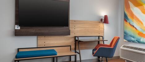 Una habitación moderna con un televisor de pantalla plana grande montado en la pared, un escritorio de madera con una silla, un banco con cojín y un cuadro abstracto colorido en la pared.